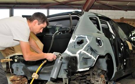 Mechanic performing vehicle body repair in a workshop, focusing on structural damage assessment and preparation for paint at Barry's Paint Shop.