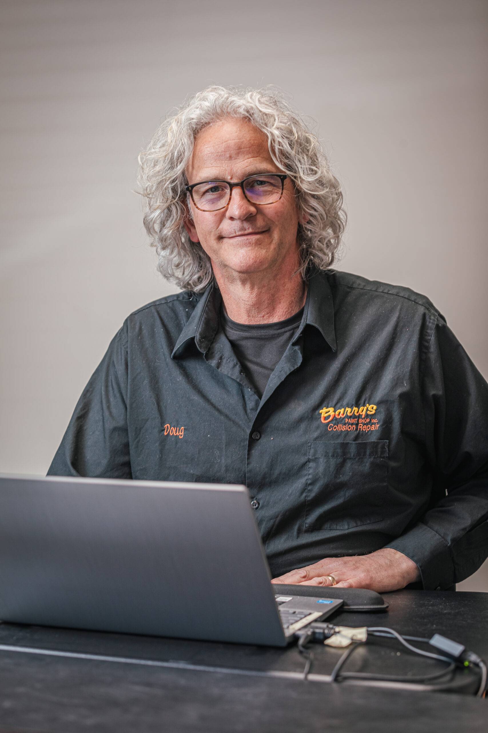 Doug Witmer, partner at Barry's Paint Shop, wearing a black shirt with the company logo, seated at a desk with a laptop, smiling, emphasizing commitment to quality automotive repair services.