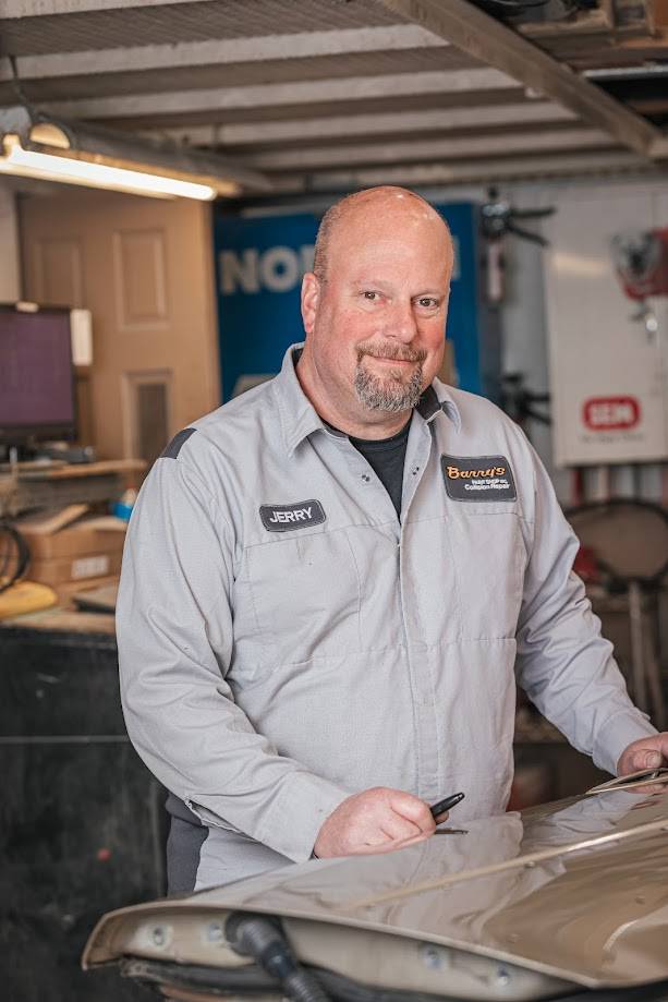 Jerry Tomalis, body technician at Barry's Paint Shop, standing in the workshop, wearing a gray uniform with name tag, holding a pen, emphasizing quality automotive repair services.