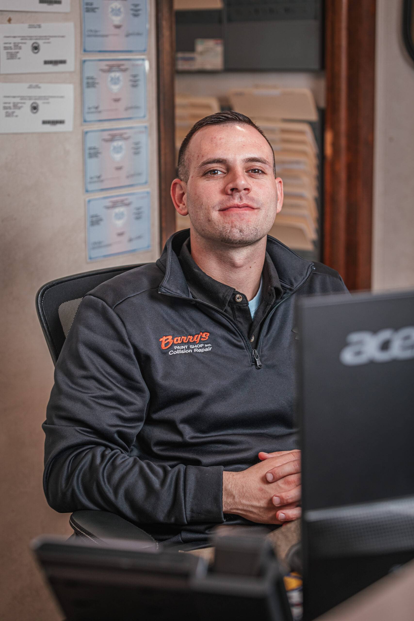 Mason Tillman, team member at Barry's Paint Shop, wearing a branded jacket, seated at a desk with visible automotive repair documents and a computer monitor, representing the company's commitment to quality service and customer satisfaction.