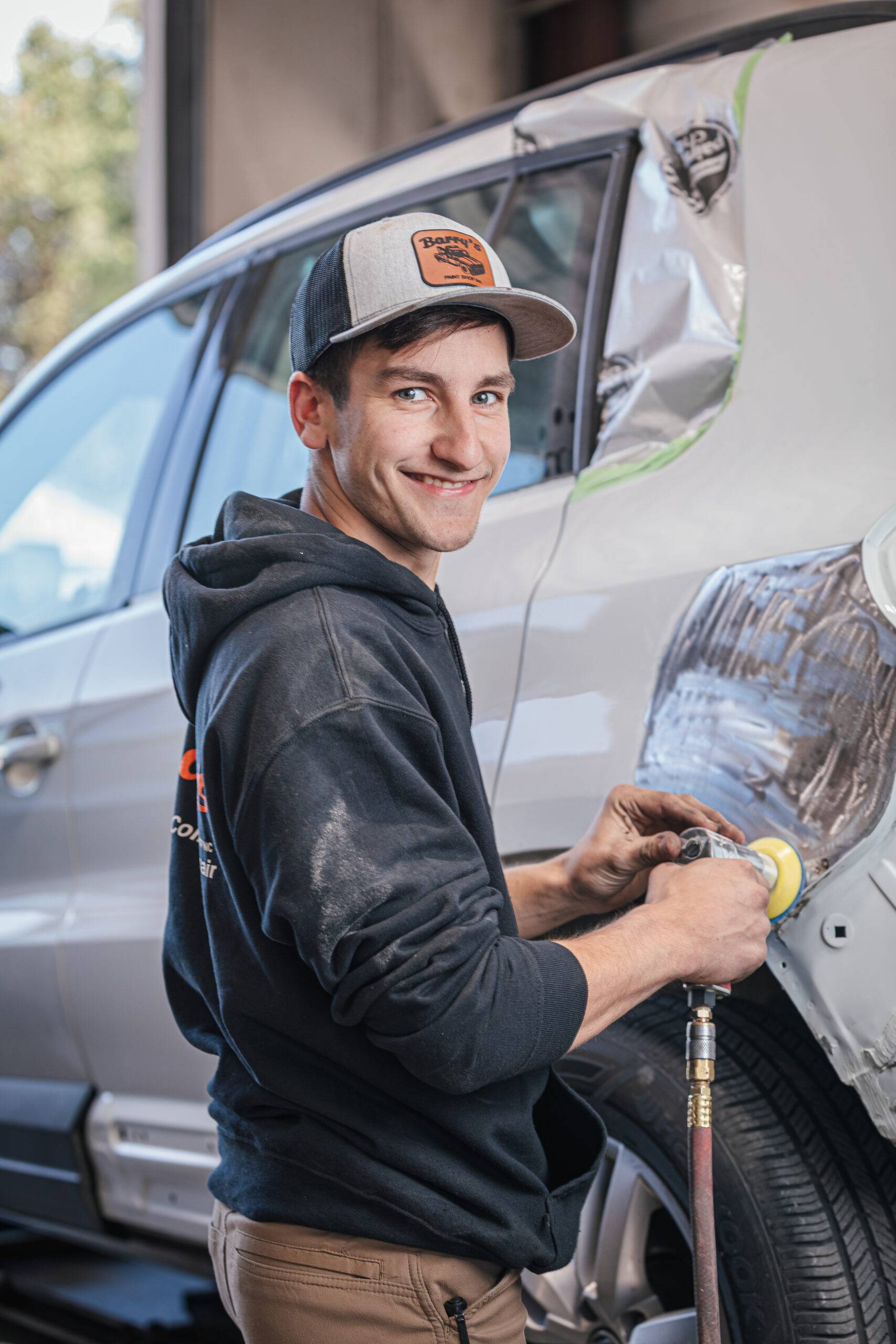 Nate Burkholder, painter at Barry's Paint Shop, working on vehicle repair with a polisher, wearing a branded cap and hoodie, showcasing expertise in automotive services.