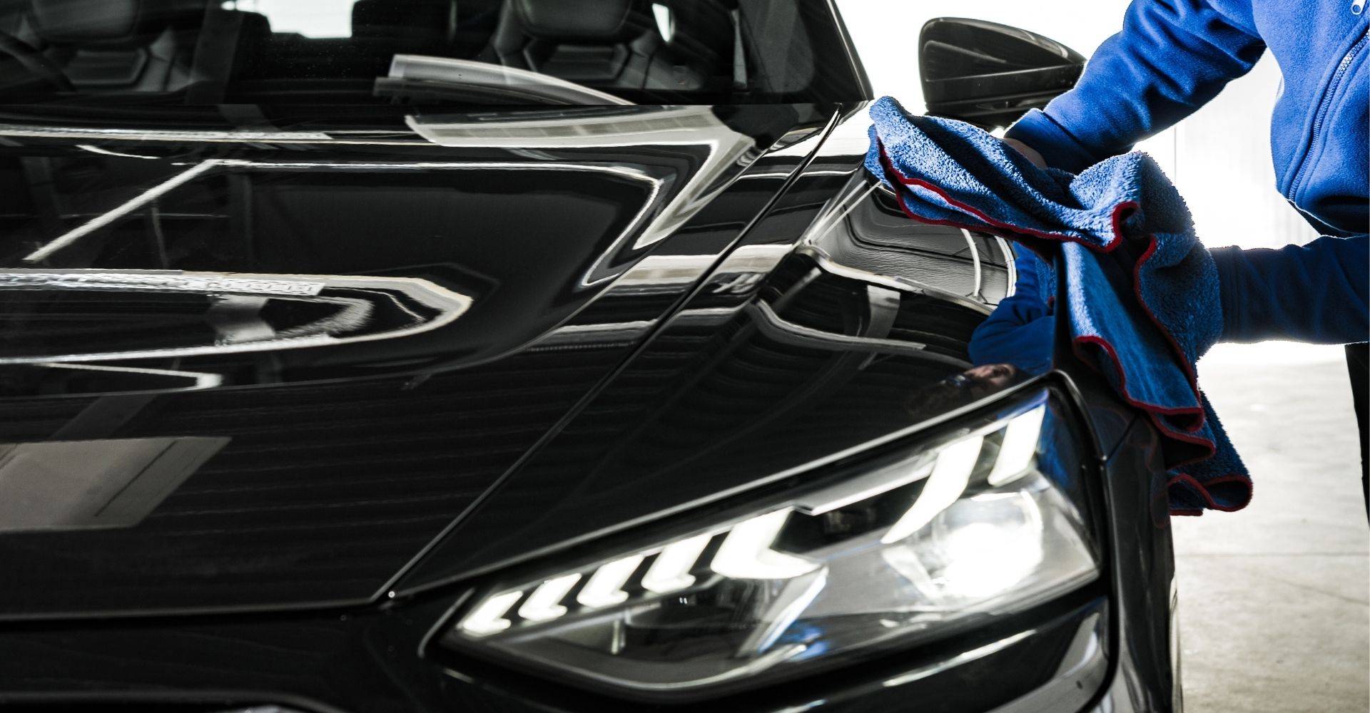A man is cleaning the hood of a black car with a cloth, focusing on removing dirt and grime.