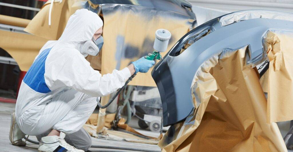 A person wearing protective gear and a mask spray paints a car bumper in a workshop.