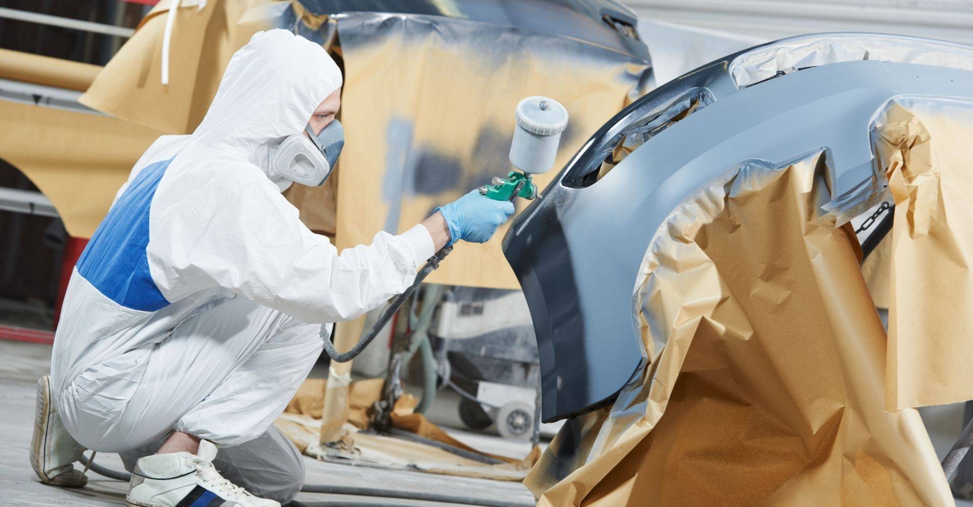 Person in protective gear spray painting a car bumper in an auto body workshop, highlighting automotive painting techniques relevant to collision repair.