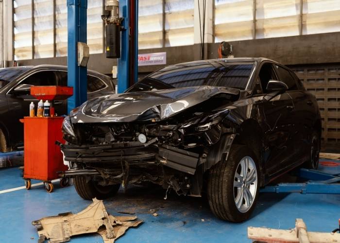 A black car with a severely damaged front end sits in a garage, supported by a blue lift