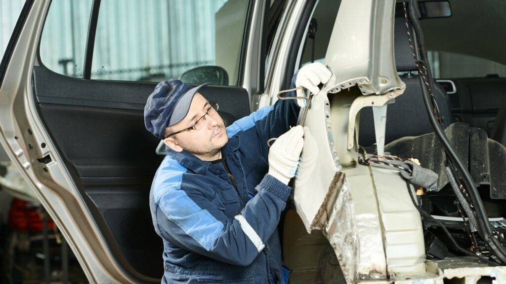 A man in a blue uniform repairs a car, focused on the engine under the hood.